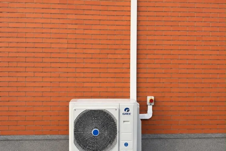 Unité extérieure de pompe à chaleur posée au pied d'un mur en briques, avec son interrupteur de coupure de sécurité