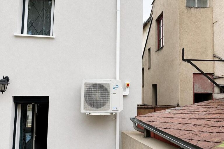 Unité extérieure de pompe à chaleur fixée en façade sur supports muraux, à côté de son interrupteur de coupure
