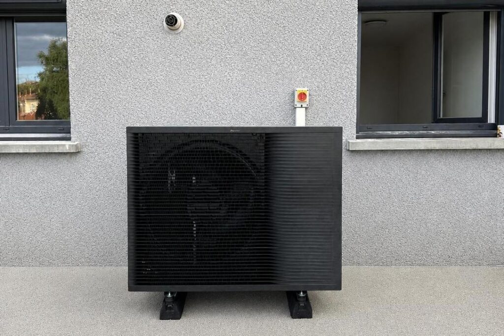 Pompe à chaleur air/eau gris anthracite posée sur plots antivibratoires au pied d'une façade, entre deux fenêtres