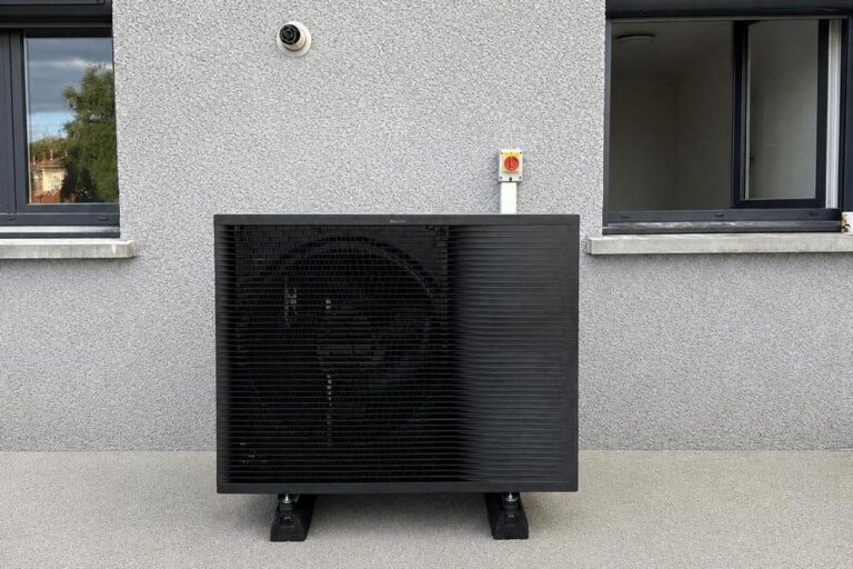 Pompe à chaleur air/eau gris anthracite posée sur plots antivibratoires au pied d'une façade, entre deux fenêtres