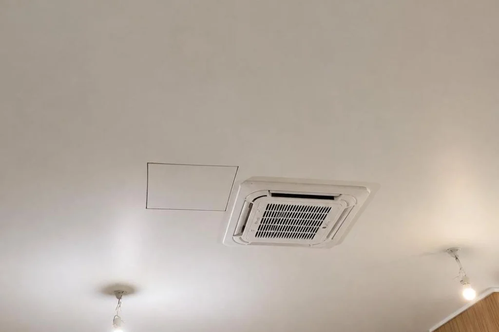 Unité intérieure de climatisation de type cassette encastrée dans un plafond blanc, à côté d'une trappe de visite