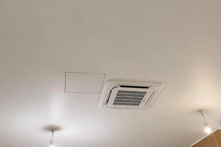 Unité intérieure de climatisation de type cassette encastrée dans un plafond blanc, à côté d'une trappe de visite